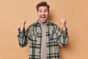 Horizontal shot of upbeat handsome man cleches fists celebrates achievement exclaims loudly wears...