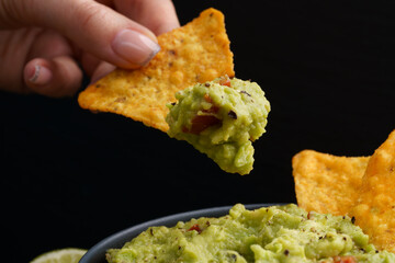 Hand with tasty tortilla chips or nachos with fresh homemade guacamole dip sauce on black background