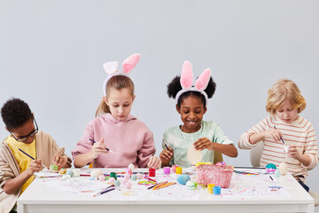 Minimal portrait of diverse group of children painting Easter eggs and wearing bunny ears while...
