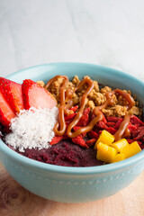 acai bowl with strawberries, granola, mango, coconut, peanut butter , green marble background, wood plate, blue bowl.