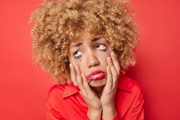 Upset crying woman with curly bushy hair has frustrated expression keeps hands on face looks with despair has broken heart feels hurt isolated over red background. Negative human emotions concept