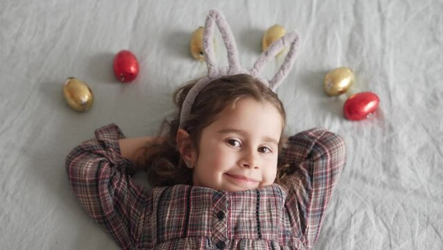Adorable little girl with curly brunette hair and a headband with bunny ears is stretched out on the bed with a grater of chocalte eggs celebrating Easter. 