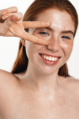 Vertical shot of happy redhead chubby girl with freckles, clear healthy facial skin, showering, cleansing her face and smiling, showing peace sign near eyes, white background