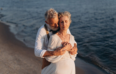 Romantic senior couple is embracing on seashore. Man hugs woman from behind.