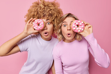 Junk food and bad eating habits concept. Surprised young women cover eyes with delicious doughnuts have unhealthy nutrition dressed casually isolated over pink background break diet. Sweet tooth