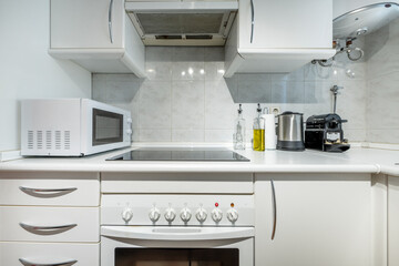 small covered kitchen with white wooden furniture, white wooden countertop, black ceramic hob, microwave, oil can and small appliances with a thermos of hot water