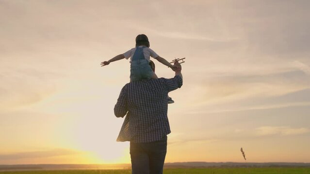 Happy family, dad, kid daughter play together with toy plane against background of spring sky. Kid, pilot sits on his dads shoulders in pilots goggles. Teamwork father, child, family dream to fly