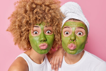 Photo of surprised two women keep lips folded apply green nourishing masks take care of complexion and skin stand next to each other isolated over pink background. Beauty and wellness concept