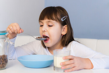 Little girl eats chocolate corn balls with milk sitting at white table. Child have breakfast