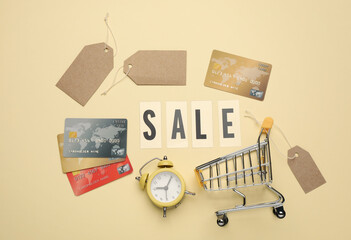 Shopping cart, tags, credit cards, alarm clock and word Sale on beige background, flat lay