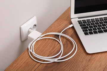 Charging laptop with power adapter in electrical socket on wooden table - obrazy, fototapety, plakaty