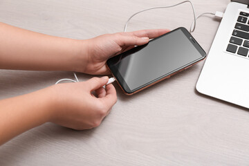 Woman connecting smartphone with charge cable to laptop on light grey table, closeup. Space for text