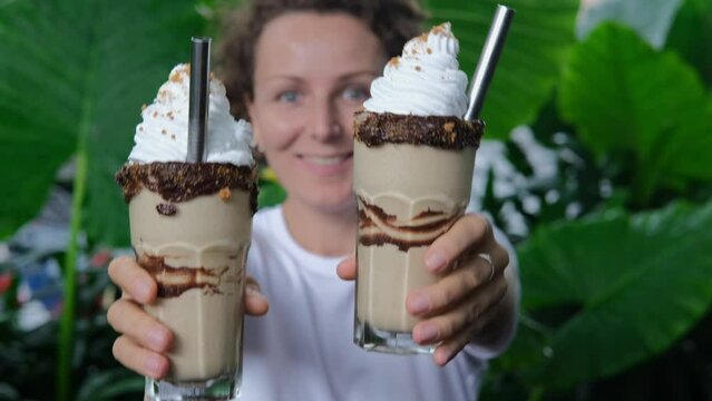 Cheerful friendly girl holding, proposing two sweet chocolate milkshakes with syrup, cream and eco metal straws