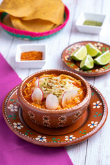 Traditional mexican food. Red pozole soup with chicken accompanied with avocado, lettuce, onion, radish, lemon, chili and crispy corn tortillas also known as tostadas on a white wooden background.