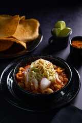 Traditional mexican food. Red pozole soup with chicken accompanied with avocado, lettuce, onion, radish, lemon, chili and crispy corn tortillas also known as tostadas on a black wooden background.