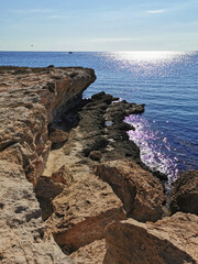 A rocky shore of long-hardened lava, a cliff, the Mediterranean Sea with a boat, against the background of a blue cloudless sky.