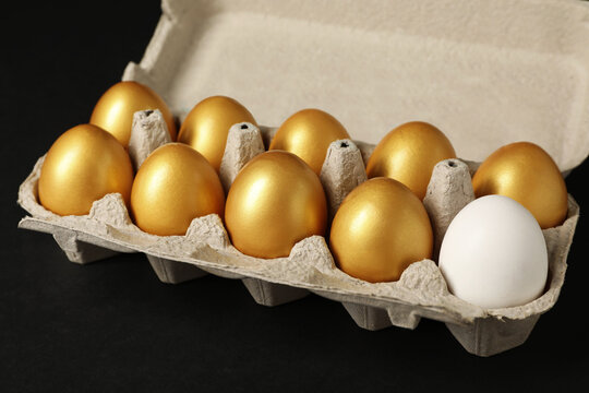 Ordinary chicken egg among golden ones in box on black background, closeup