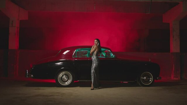 Beautiful Elegant Young Woman Standing , Dancing and Flirting near old retro classic car. Old fashioned luxury style concept . Rich fashion lifestyle . Luxury dress with fabric glitter, spangles .