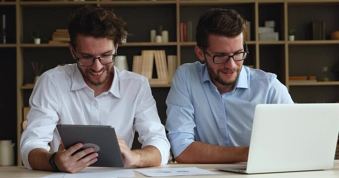 Laughing twin brothers students wear glasses sit at desk engaged in distant learning using laptop tablet discuss common task. Young men twins work on business project together enjoy creating new ideas