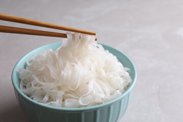 Taking rice noodles with chopsticks from bowl at light grey table, closeup
