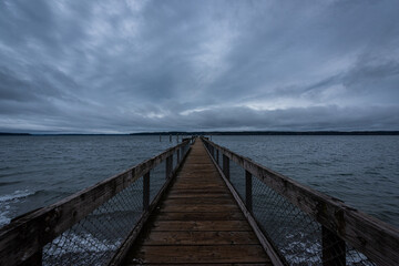Fototapeta na wymiar Long wooden pier stretching into the horizon over water on cloudy overcast blue hour morning