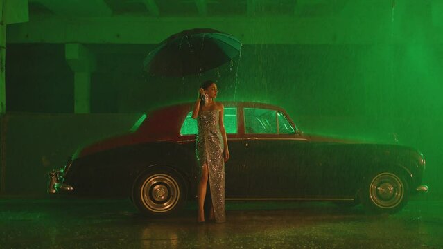 Beautiful Elegant Young Woman Standing and holding umbrella near old retro classic car. Girl with umbrella stands in street under the rain . Old fashioned luxury style concept . Autumn rainy day