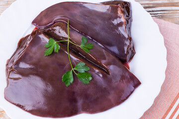 Raw liver on a white plate on a wooden background. Beef liver, top view. Offal in its raw form.