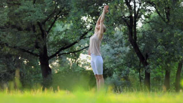 Young woman doing yoga practice or fitness while standing in green summer park spbi. 4k video Attractive athletic female does asana or pose and raises hands to sky, stands on grass and stretches her