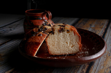 A clay or mud Mexican hand made plate, with sweet bread and raisins on top and a steaming cup of coffee or hot chocolate cocoa in the background. Horizontal dark food photography