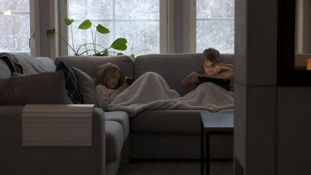 Children sit on couch with pad and phone, girl smiles into camera