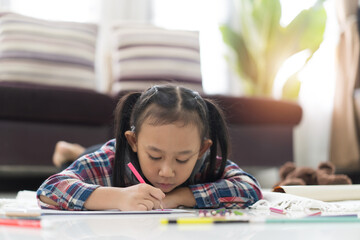 Happy cute asian little girl drawing using colorful pencils while lying on the floor at home