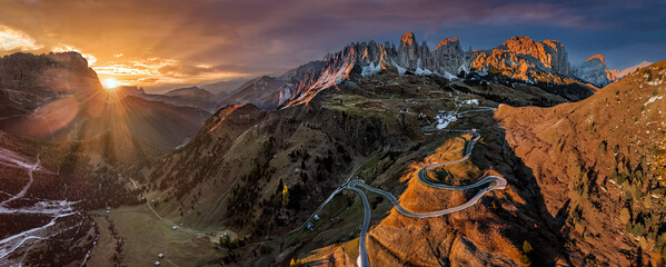 Gardena Pass, Italy - Aerial panoramic view of the curvy roads of the famous Gardena Pass (Passo Gardena) with the peaks of Pizes de Cir, the Italian Dolomites and a warm sunset at autumn season