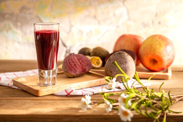 A glass of red flavor mixed fruit juice serves in a tray with piece of beetroot, passion fruit, and apple on wooden table over colorful wall background. Eatable recipe health herb vegan drink concept