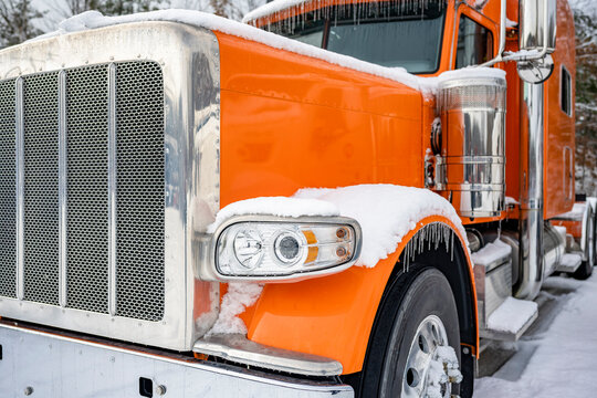Powerful classic orange big rig semi truck with extended cab standing on the winter parking lot with snow and ice waiting for the next load