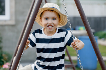 Funny little boy in a hat and striped T-shirt is having fun with a chain swing on an outdoor playground. Child swings on a sunny summer day. Active recreation with children.