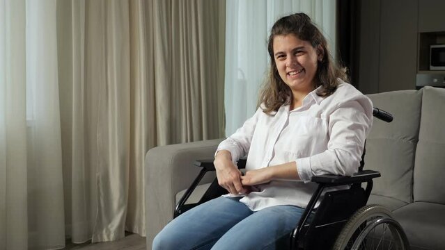 Young disabled woman with infantile cerebral paralysis in white shirt smiles sitting in wheelchair near stylish sofa in living room at home.