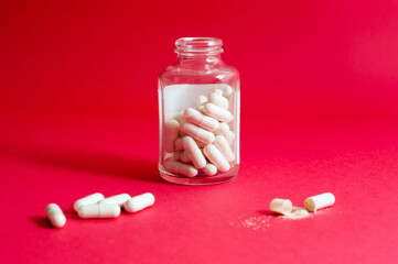 an open glass jar with pill capsules stands on a red background, part of the tablets lies on the background, one capsule is open and part of the medicine has spilled out of it.