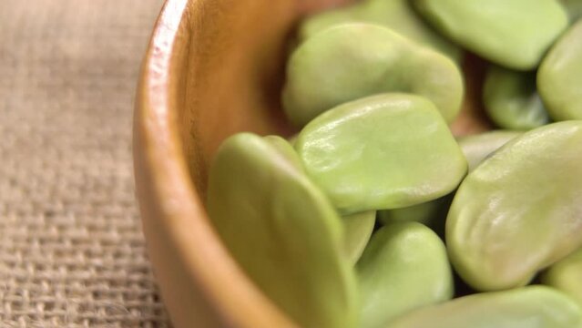 Falling green dry broad or fava beans in wooden bowl on rustic burlap. Macro. Rotation. Legume family. Slow motion