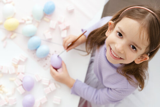 little girl with hearing aid paints easter eggs purple, happy child paints easter eggs on white wooden table