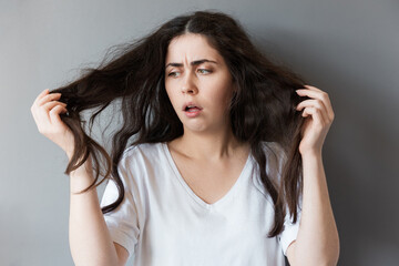 A young Caucasian woman looks at her disheveled long dark hair in bewilderment. Gray background....