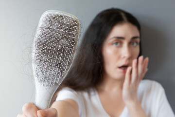 Blurred portrait of a shocked Caucasian young woman shows her hairbrush with losing hair. Gray...