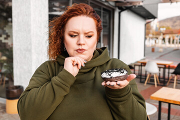 Portrait of plus sized thougthful woman holds a chocolate donut at hand. The concept of diet and...