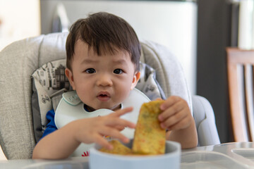 Adorable joyful and Happy 1 years old Chinese baby boy having food while sit on baby chair