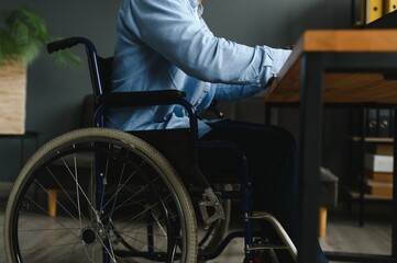 Obraz premium Young man with special needs in casual clothes working on wireless laptop. Male freelancer working from home while sitting in wheelchair.