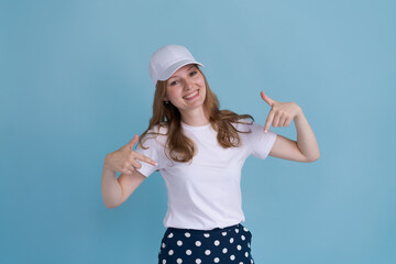 Portrait cheerful girl straight fingers herself on in white cap on blue background. Caucasian successful young woman shows that everything is fine with her. Advertising concept