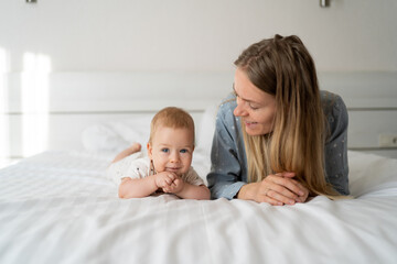 mother with baby six months old in the bedroom. Young happy mum lies next to her adorable baby daughter playing in white bed enjoying sunny morning