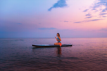 A young smiling woman poses sitting on a sup board with paddle. Sunset in the background. Copy...