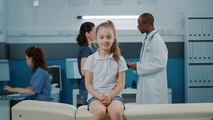 Obraz premium Portrait of small child waiting to receive medical advice from doctor in office. Little girl sitting on bed in cabinet, preparing for consultation appointment with professional pediatrician.