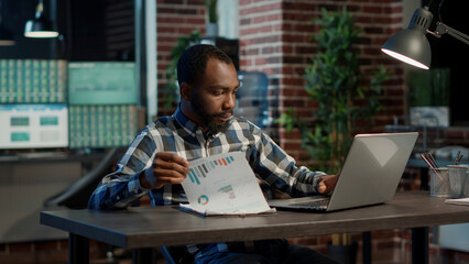 Sales consultant analyzing financial charts on papers and laptop, using statistics to plan new...