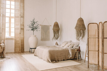 Spacious, bright bedroom with white walls and a large bed in warm bohho tones. Straw chandeliers, a large decorative vase, wooden shutters on the windows.